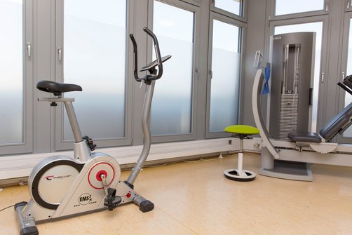 Trainingsraum mit Fahrradergometer, Zugapparat und grünem Hocker.