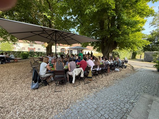 Große Gruppe von Kolleg:innen sitzt gemeinsam an langen Tischen unter Bäumen im Garten des Hofguts.
