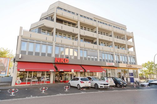 Straßenansicht des Ärztehaus-Gebäudes mit Bekleidungsgeschäft, Versicherung und Bäckerei im Erdgeschoss, parkende Autos davor.